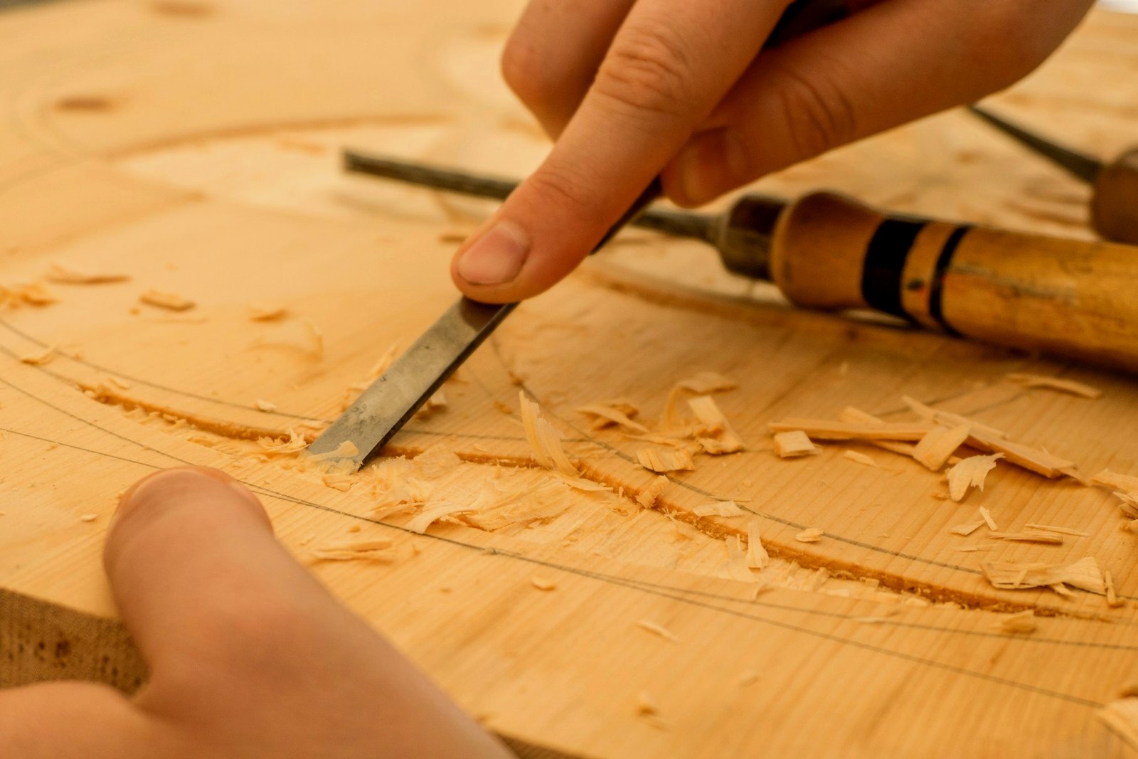Nahaufnahme einer Werkbank: eine Hand führt einen Stechbeitel entlang einer angerissenen Linie durch helles Holz, Späne liegen ringsum, daneben zwei Stecheisen mit Holzgriff